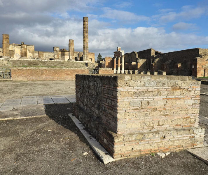 The ruins of the Temple of Jupiter at Pompeii, with weathered columns, a brick podium, and a partially intact structure under a bright sky with scattered clouds.