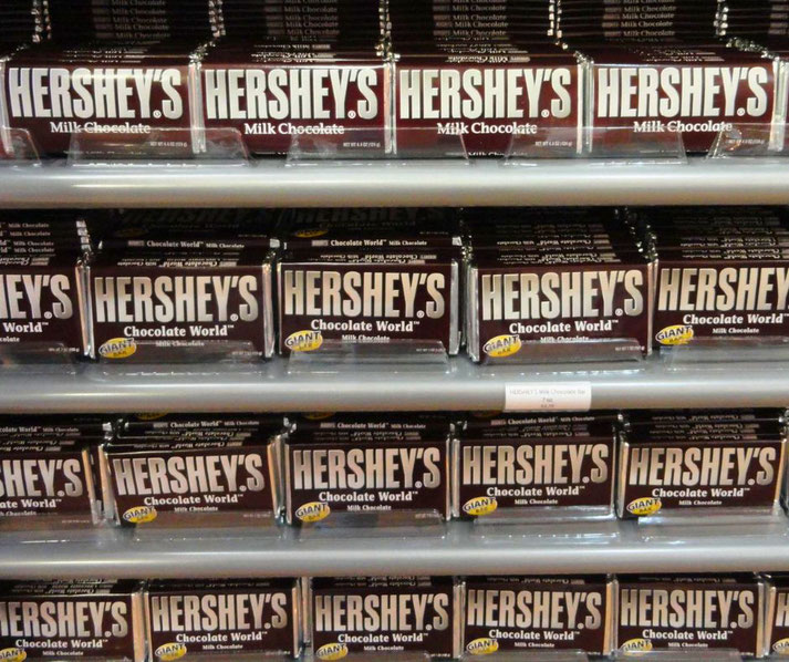 A store shelf filled with large Hershey's Milk Chocolate bars, neatly stacked in rows. Some are labeled "Chocolate World" and "GIANT," emphasizing their size.
