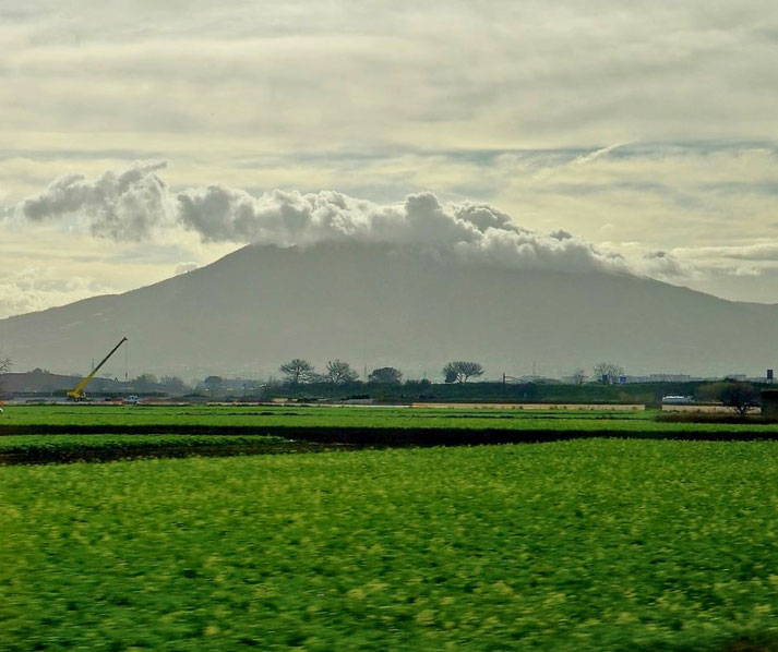 A cloud-covered mountain rises behind vibrant green fields, with a crane visible in the distance under a hazy sky.