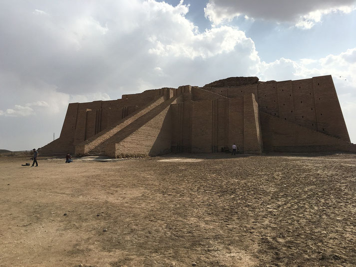 The Ziggurat of Ur in modern-day Iraq, an ancient Mesopotamian structure with grand staircases leading to a temple platform. The sky is partly cloudy, and a few visitors are present.