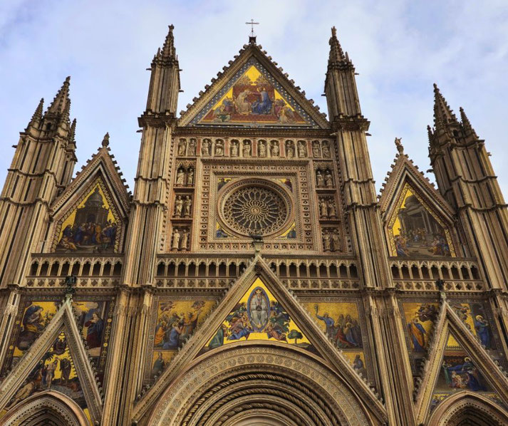 The ornate Gothic facade of the Orvieto Cathedral (Cattedrale di Santa Maria Assunta) in Italy. It features intricate sculptures, a large rose window, and vibrant golden mosaics.