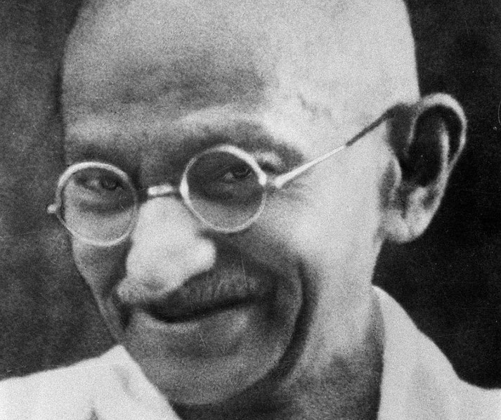 Close-up black-and-white photo of an elderly man with round glasses, a mustache, and a gentle smile, wearing a simple white garment.