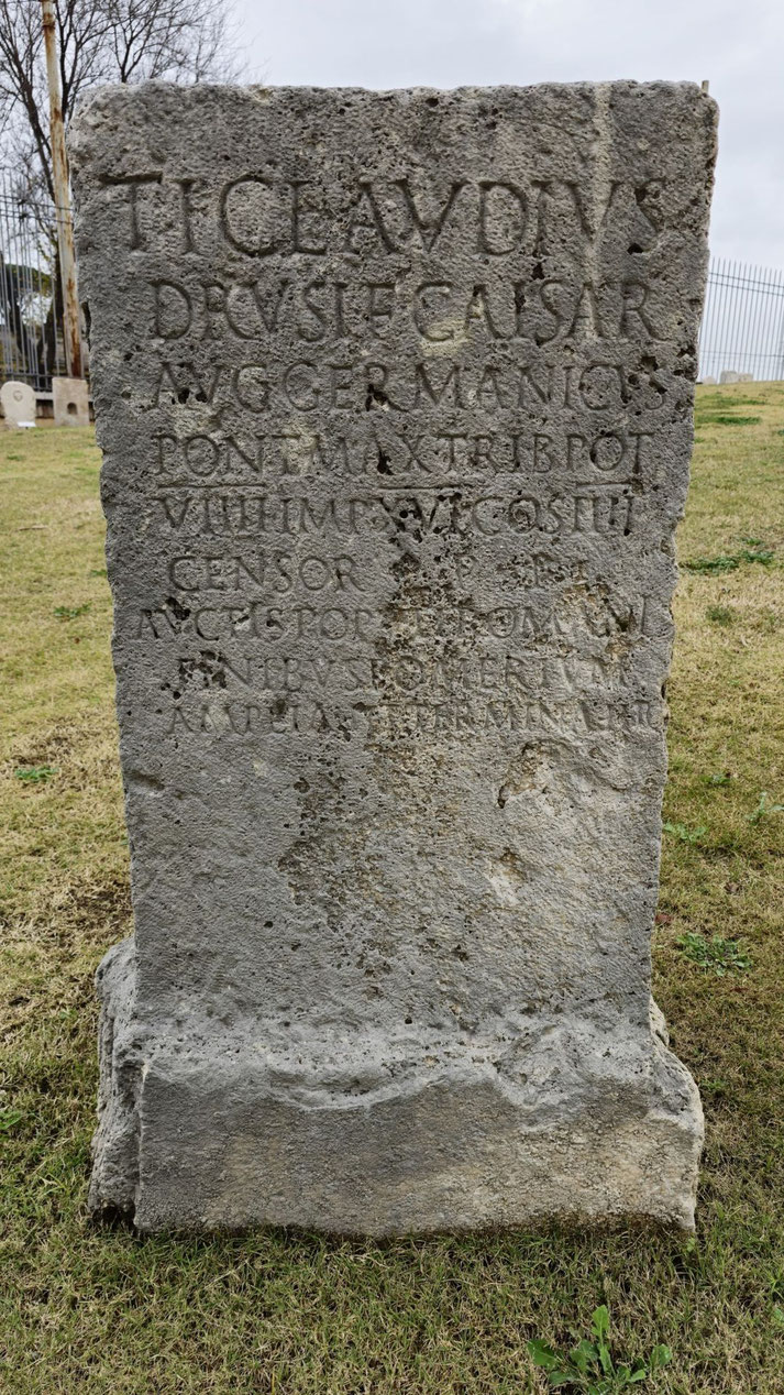 Weathered Roman stone slab with Latin inscription honoring Emperor Tiberius Claudius. Set outdoors on grass with fencing and ruins in the background.