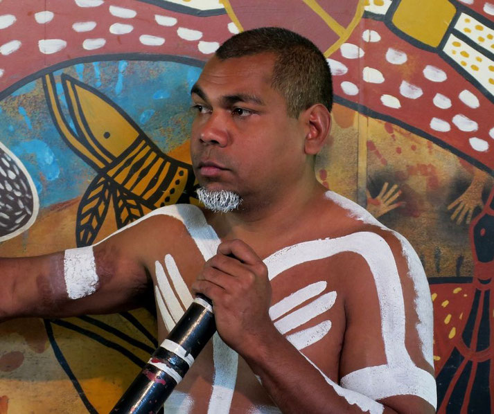 A man with traditional white body paint holds a didgeridoo, standing before a vibrant Indigenous artwork.