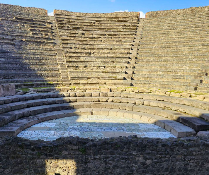 Odeon of Pompeii, a well-preserved ancient theater with stone seating arranged in a semi-circle, a central mosaic floor, and sunny and shadowed sections under a clear blue sky.