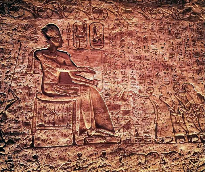 Ancient Egyptian wall carving shows a seated figure with extended arms facing a group of standing figures, surrounded by dense hieroglyphic text.