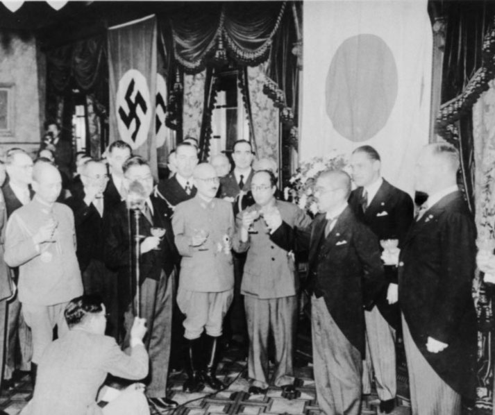 Japanese and German officials toast at a diplomatic event during World War II, with Nazi and Japanese flags displayed prominently in the background.