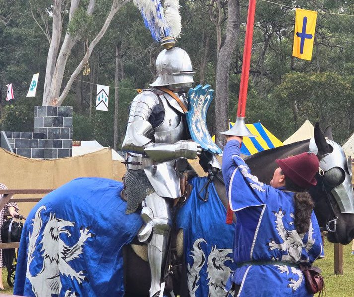 A knight in full plate armor on a caparisoned horse at a jousting event.