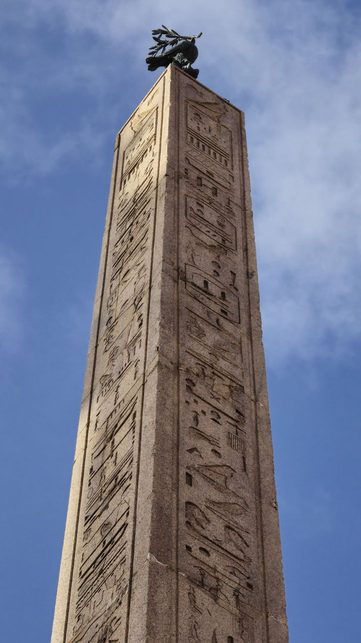 Tall ancient obelisk with Egyptian hieroglyphs, topped by a bronze sculpture, rising sharply against a blue sky.