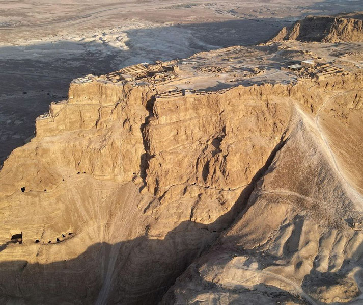 Aerial view of a desert fortress atop a steep plateau with ruins, surrounded by barren cliffs and winding paths below.