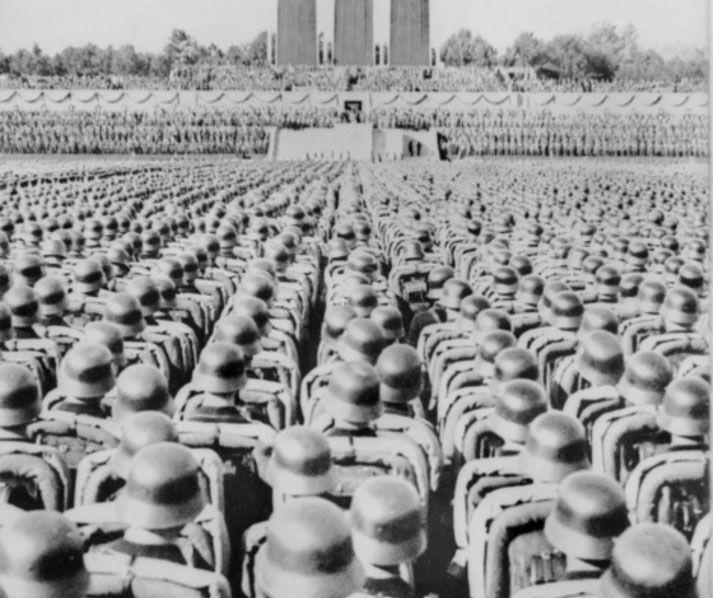 Large formation of German soldiers in uniform and helmets assembled during a Nazi rally in the 1930s, viewed from the rear.