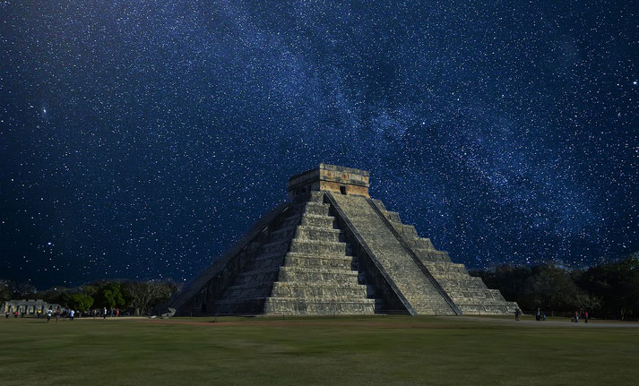 El Castillo at Chichén Itzá under a star-filled night sky. The ancient Mayan pyramid stands illuminated against the dark backdrop, surrounded by lush greenery and visitors in the distance.