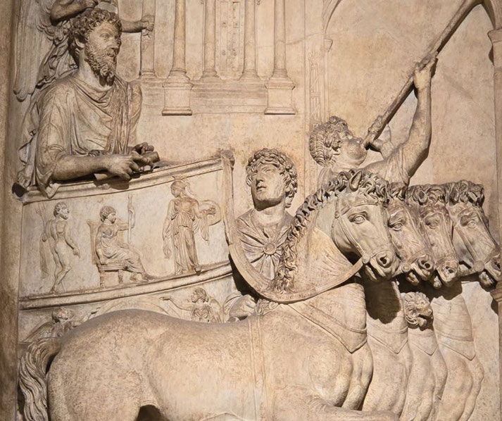 Elaborate stone relief of a Roman chariot procession with four horses, a horn-blower, and figures holding scrolls.