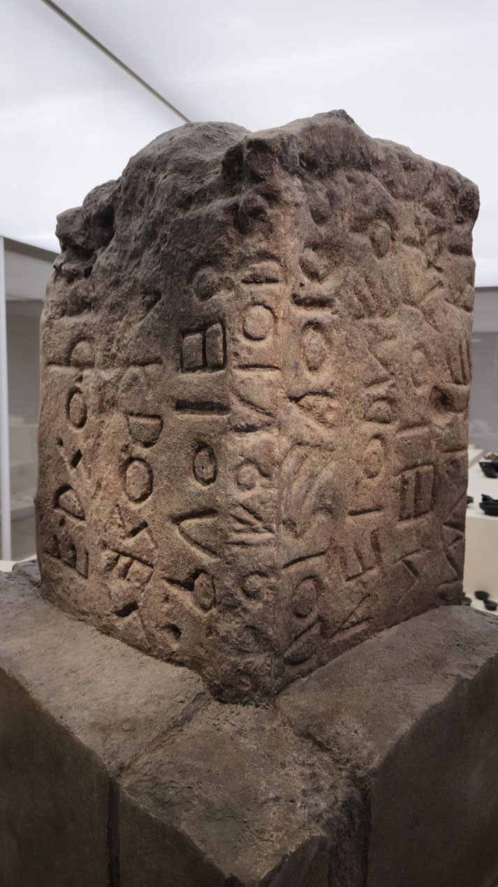 Ancient stone pillar carved with geometric and symbolic script, possibly an early form of writing or proto-alphabet, displayed in a museum setting.