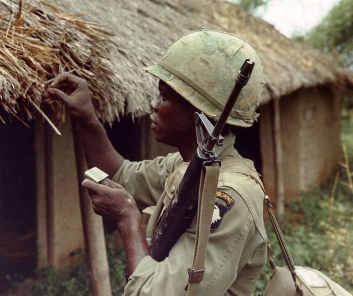 A U.S. paratrooper in uniform lights a match near a thatched hut, holding a rifle slung over his shoulder.