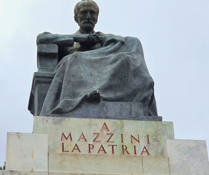 Bronze statue of a seated Giuseppe Mazzini, draped in a cloak, atop a marble pedestal inscribed with “A MAZZINI LA PATRIA.”