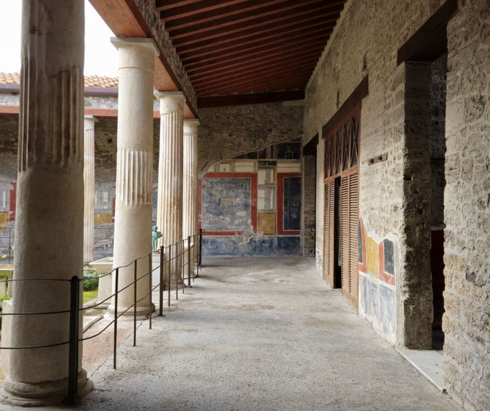 Stone corridor with columns and preserved frescoes in an ancient Roman house, with a garden visible through the colonnade.
