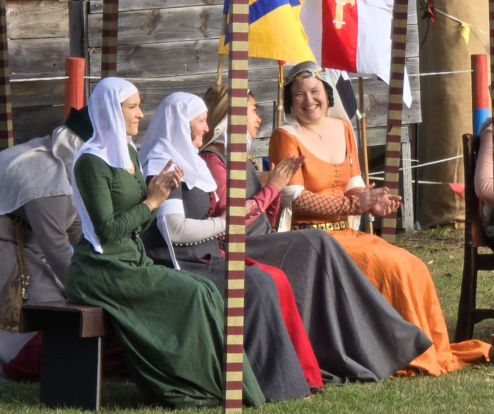A group of women in medieval attire, including colorful dresses and head coverings, sit and clap during an outdoor event. Their cheerful expressions and flags in the background suggest a festive gathering.