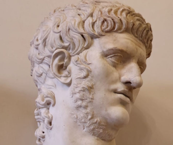 Marble bust of a man with curled hair, sideburns, and a faint mustache, shown in a tilted pose with a confident expression, likely depicting the Roman Emperor Nero.