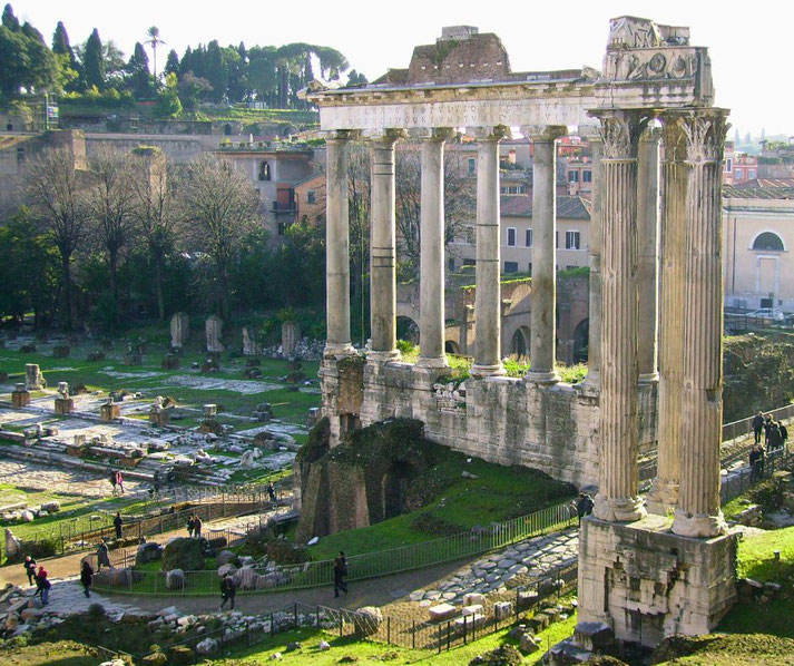 Ruins of ancient Roman columns and foundations, part of a historic temple structure, surrounded by grassy paths and visitors in the Roman Forum.