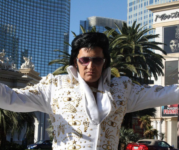 Man dressed as Elvis Presley in a white rhinestone jumpsuit and sunglasses, posing with arms outstretched on a sunny day in Las Vegas.