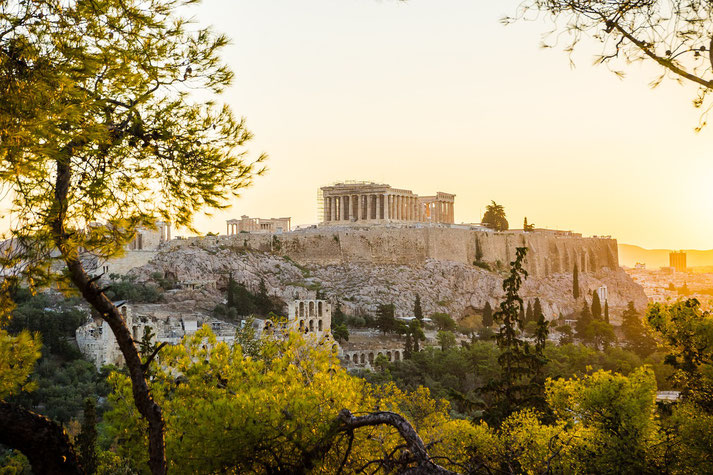 Acropolis in Athens, Greece, featuring the Parthenon atop a rocky hill surrounded by lush greenery, with a golden sunset casting a warm glow over the ancient ruins.