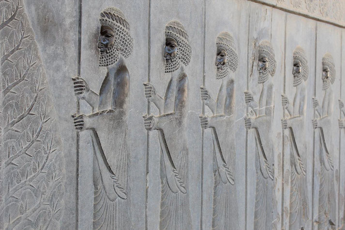 Ancient stone relief showing a row of bearded Persian guards holding spears, carved in profile with stylized robes and headdresses.