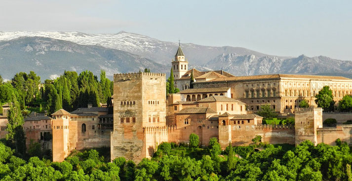 Alhambra in Granada, Spain, a stunning Moorish palace and fortress surrounded by lush greenery, with the Sierra Nevada mountains providing a majestic backdrop under a clear sky.