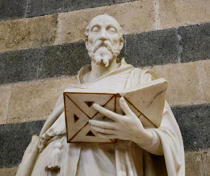A marble statue of a bearded man holding an open book with geometric carvings, set against a striped stone wall, gazes forward with a composed expression.