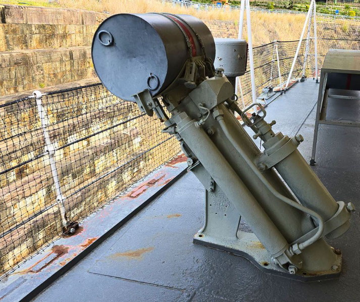 A depth charge launcher is mounted on a ship deck, holding a single charge with a red band, aimed outward beside a safety net.