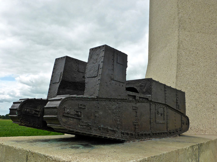A metal sculpture of a WWI tank with twin turrets, riveted armor, and tracks, mounted on a stone base near a large monument.