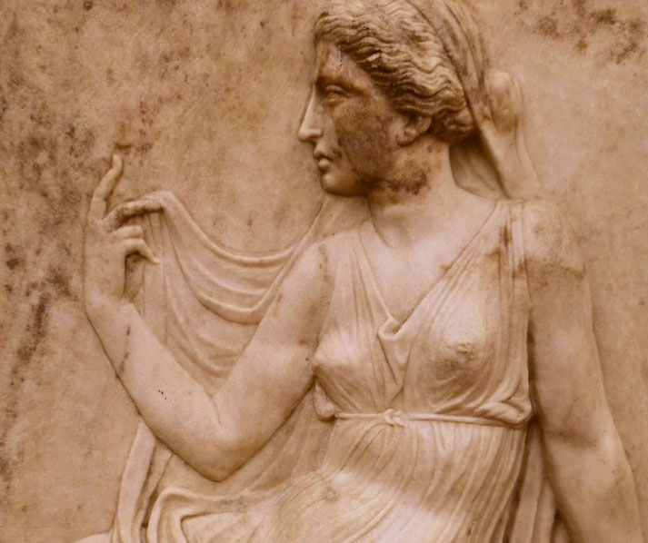 Marble relief of a woman in a flowing gown, gracefully lifting her draped veil with delicate fingers, showing calm elegance and classical style.