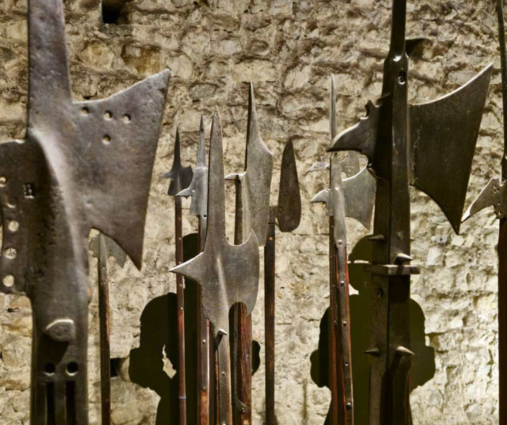 A collection of medieval polearms, including halberds, displayed against a stone wall. The weapons have sharp, intricate metal blades mounted on wooden shafts.
