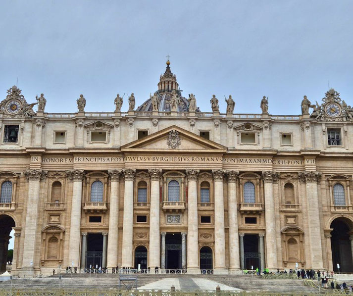 The grand facade of St. Peter's Basilica in Rome, featuring massive columns, statues of saints, Latin inscriptions, and the iconic dome rising in the background.