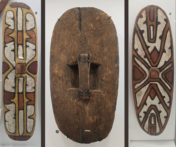 A triptych of Indigenous Australian shields. The outer shields feature intricate geometric and organic patterns in earthy tones, while the center shield is plain.