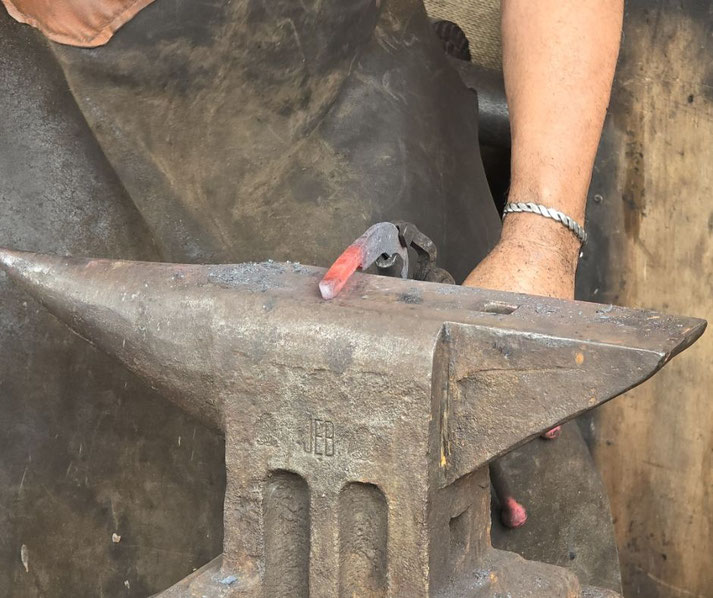 A blacksmith's anvil with a heated, partially forged metal piece resting on it. A worker, wearing a leather apron and metal bracelet, is engaged in metalworking.