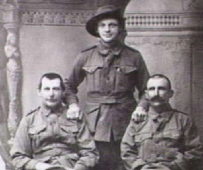 Three soldiers in vintage military uniforms pose for a studio portrait; two sit while one stands behind with hands resting on their shoulders.