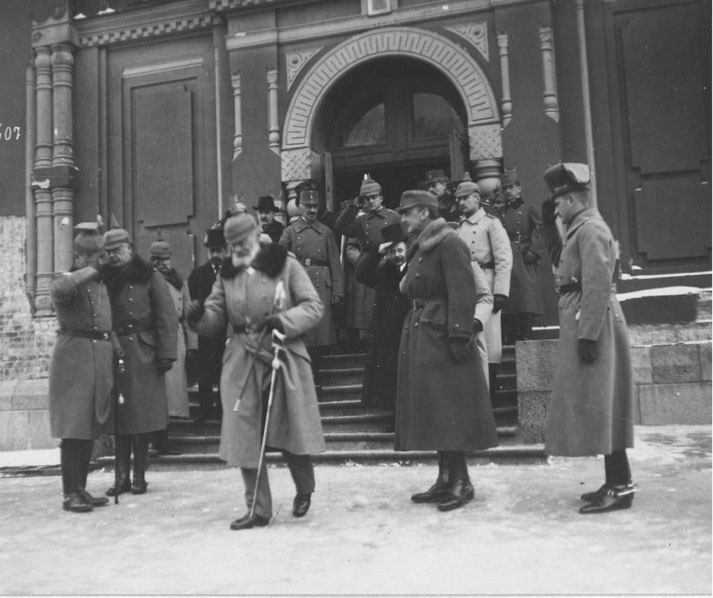 Black-and-white photo of military officers and civilians outside a large railway station building. An elderly bearded man with a cane walks across the foreground while soldiers stand at attention.