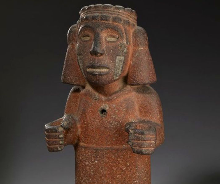 A grey‑stone Aztec sculpture of the water goddess Chalchiuhtlicue, shown kneeling, wearing a broad tasselled headdress and serpentine belt, with red pigment traces.