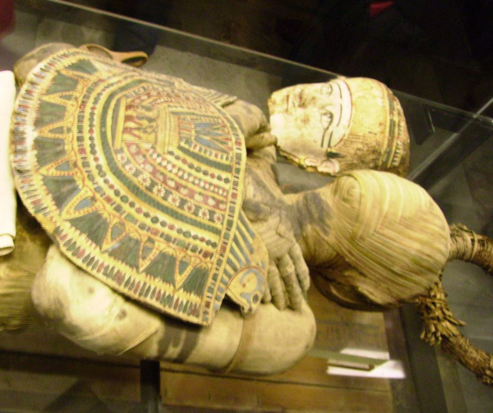 An ancient Egyptian mummy with intricate wrappings, a decorative chest plate, and a painted funerary mask. The mummy is displayed in a glass case, preserving its detailed burial adornments.