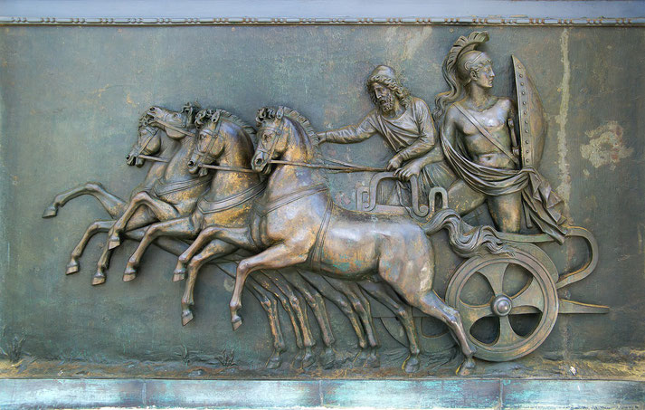 Bronze relief depicts four galloping horses pulling a chariot with two ancient figures, one armored with a helmet and shield, the other holding reins.