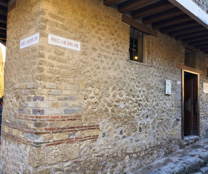 Lupanar of Pompeii, a brothel with stone walls, a wooden doorway, barred windows, and signs marked "Reg. VII. Ins. XII," representing its location in the ancient city.