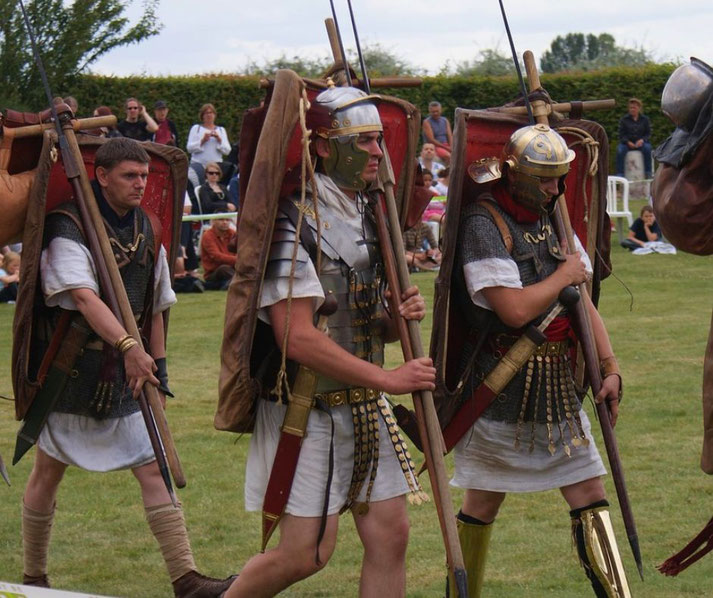 Reenactors dressed as Roman legionaries march in formation carrying shields, spears, and gear during a public event.