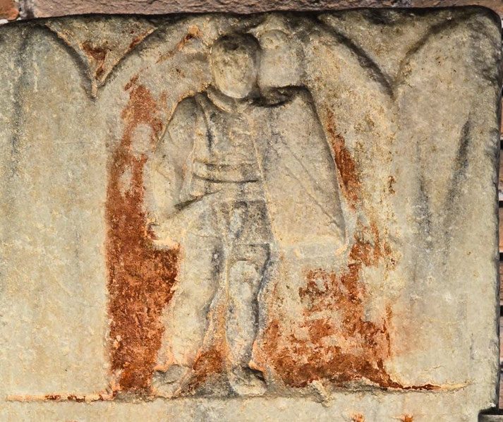 Faded stone relief of a standing figure in a cloak, partially eroded with traces of red pigment around the body and background.