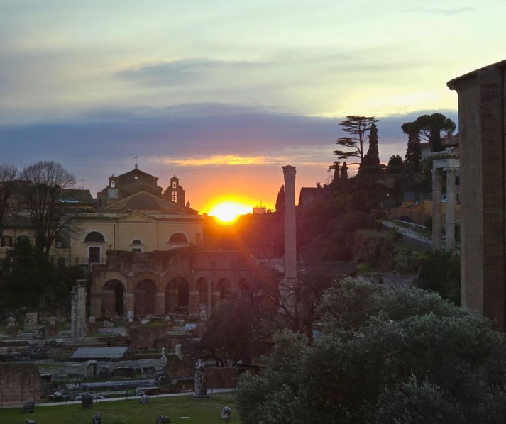 A sunset over the Roman Forum, casting a golden glow on ancient ruins, historic buildings, and towering columns.