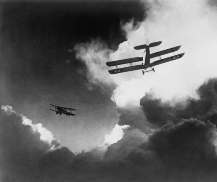 Two vintage biplanes fly through dramatic clouds, one higher and closer, the other trailing in the distance.
