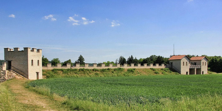 A reconstructed Roman fort with stone walls, watchtowers, and a gated entrance. The structure stands against a green field under a clear blue sky.