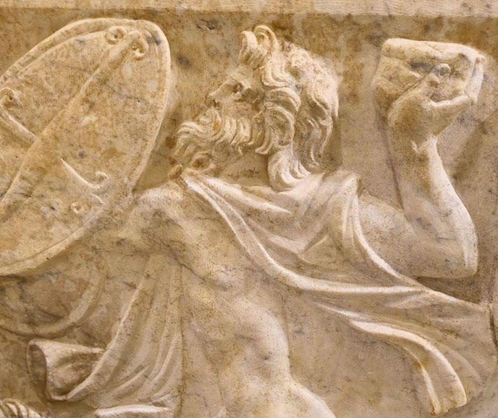 Marble relief of a bearded warrior holding a round shield and raising a stone, captured mid-action with flowing garments.