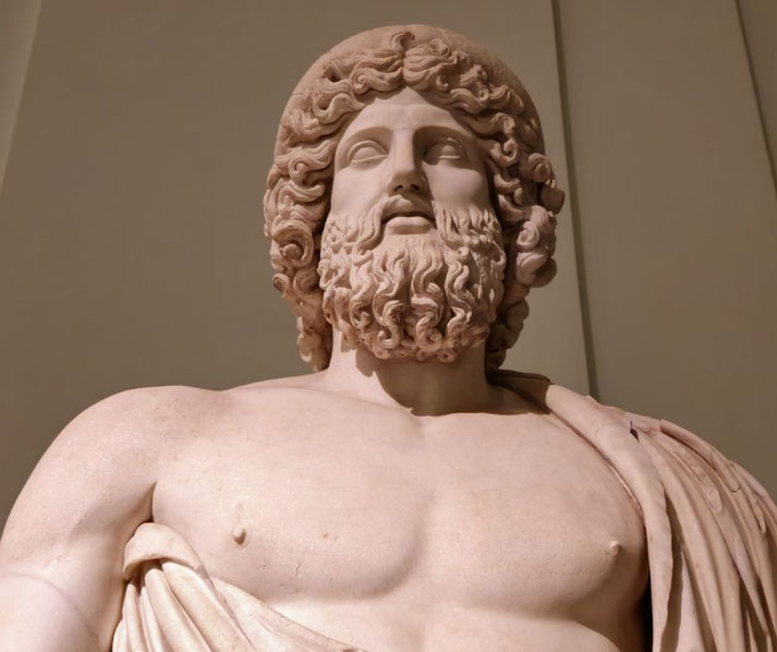 Close-up of a muscular bearded male statue with curly hair and draped clothing, gazing upward with a serene, powerful expression.