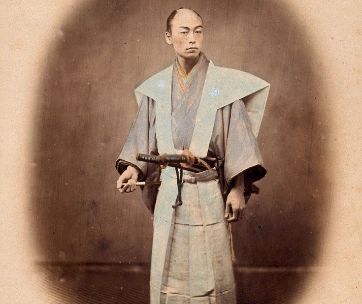 A hand-colored vintage photograph of a samurai in traditional attire, holding a sheathed sword. He wears a formal kimono with a distinctive layered collar and wide-shouldered vest.